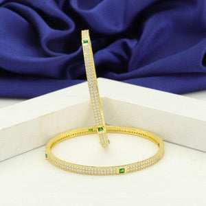 Elegant gold bangles with emerald accents and intricate detailing on a blue satin background.