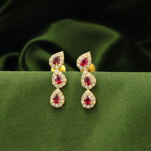 Elegant gold drop earrings with red gemstone and diamond accents displayed against a lush green fabric background.